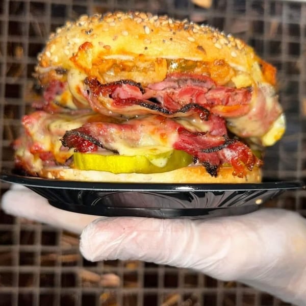 Gloved hand holding a deli sandwich with pastrami, melted cheese, and pickles on a sesame bun.