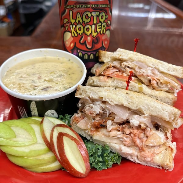 Deli sandwich with soup, apple slices, and a "Lacto Kooler" drink on a red plate.
