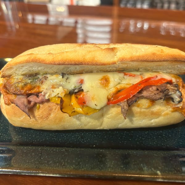 A Harvest deli sandwich with melted cheese, roasted beef, and colorful bell peppers on a dark plate.