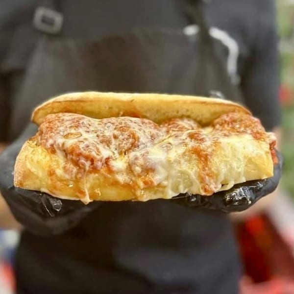 Hands in black gloves holding a Harvest deli meatball sub with melted cheese and sauce.