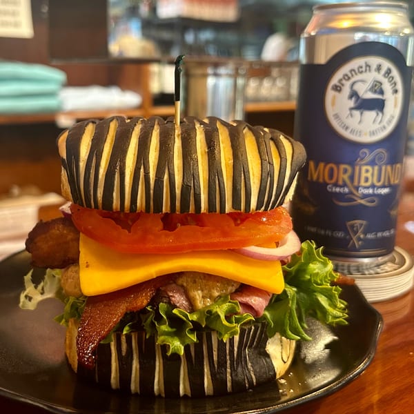 A deli sandwich with a striped bun, cheese, bacon, lettuce, tomato, and onion, next to a beer can.