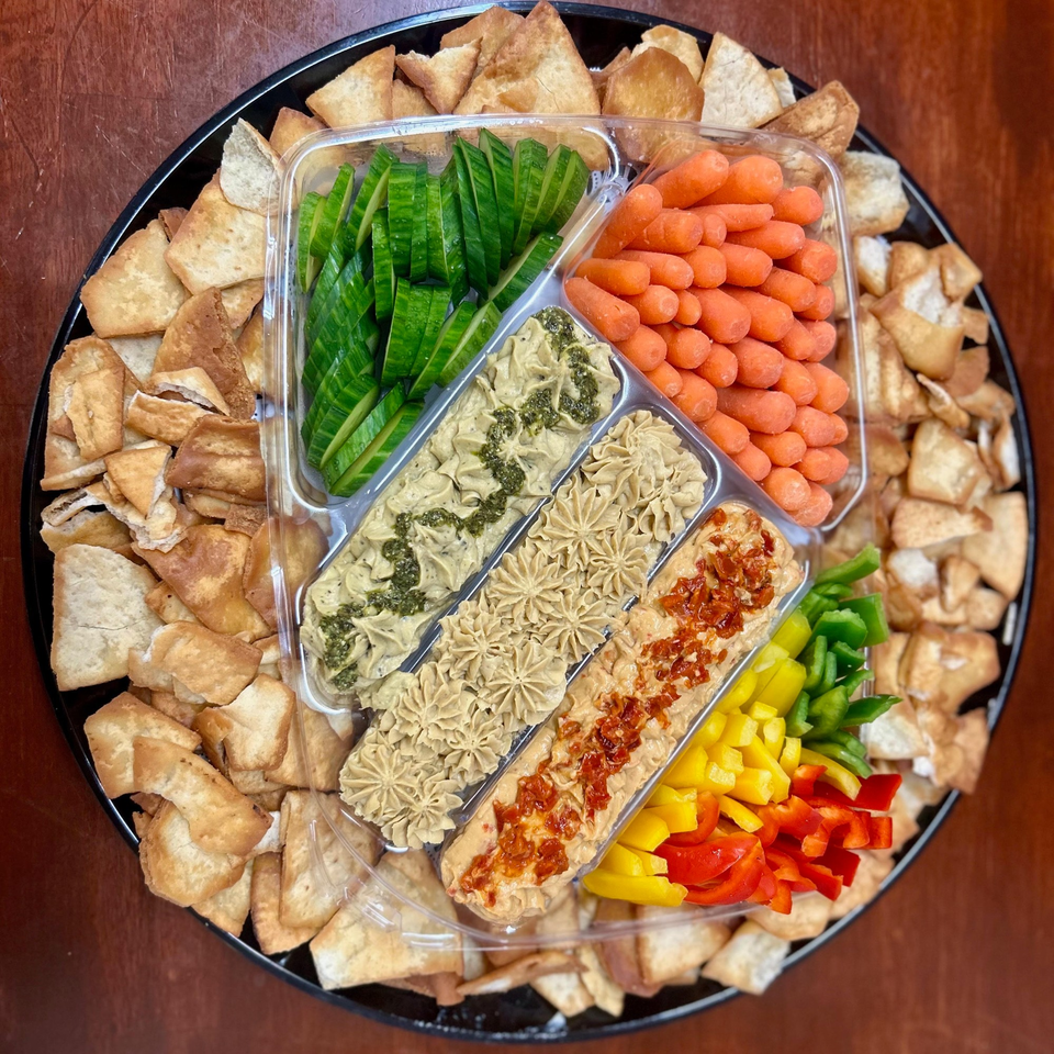A Harvest deli platter with pita chips, sliced cucumbers, baby carrots, bell peppers, and three dips.