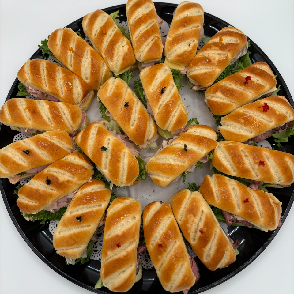 A black platter filled with Harvest deli's mini baguette sandwiches, featuring various fillings and toothpicks.